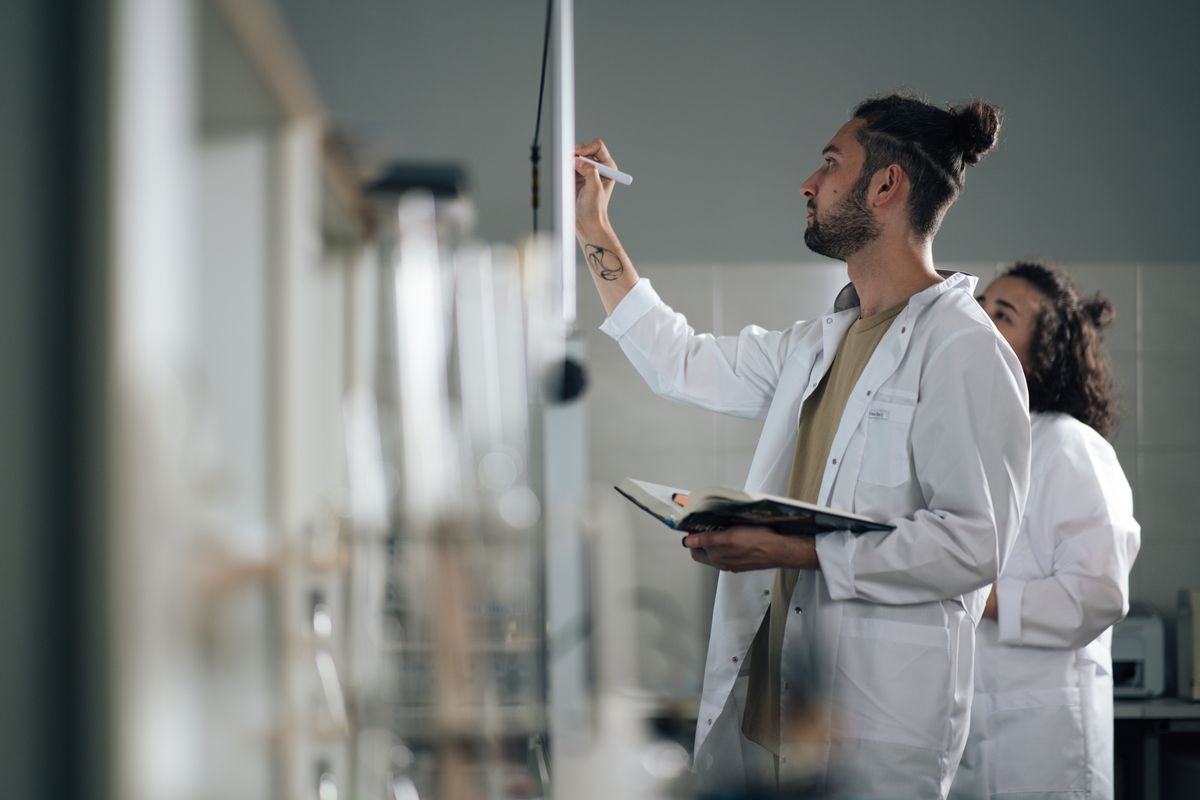 Man in a laboratory coat writing on a whiteboard while holding a notebook.