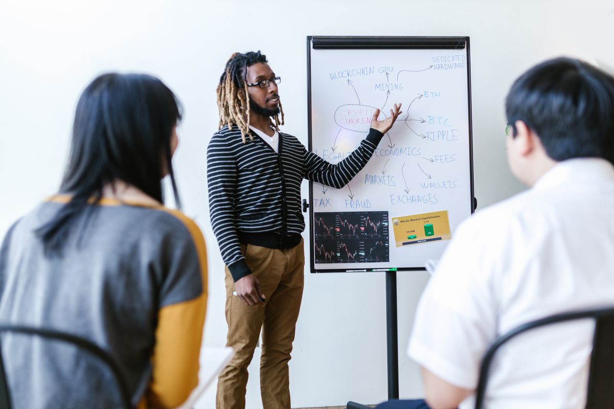 Presenter explaining a cryptocurrency diagram on a whiteboard to seated participants.