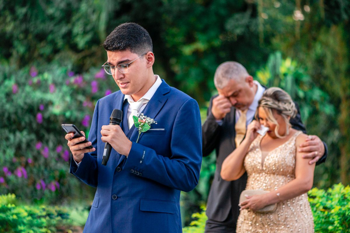 Groom in a blue suit reading from his phone and speaking into a microphone outdoors.